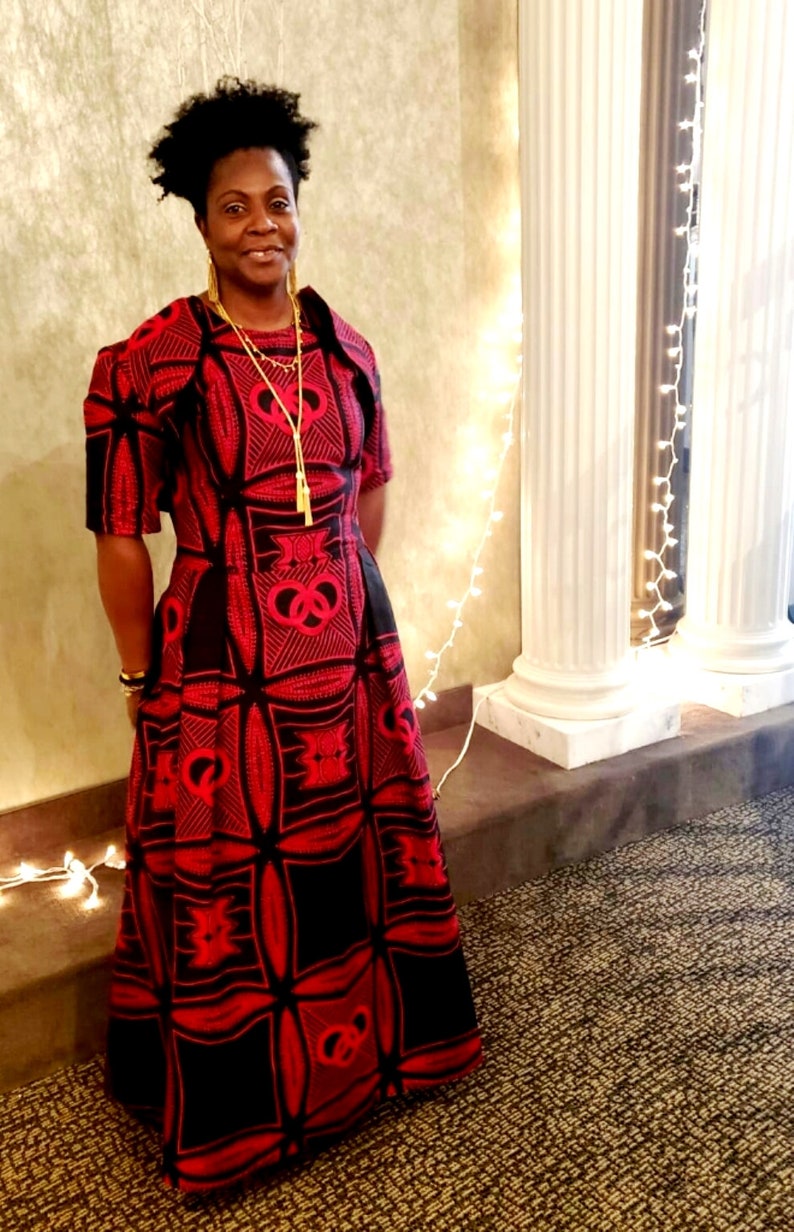 May include: A woman wearing a red and black patterned dress with a gold necklace. The dress has a fitted bodice and a full skirt. The woman is standing in front of a white wall with string lights.