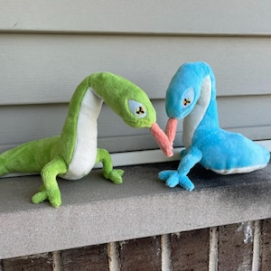 May include: Two plush lizard toys. One is green with a white belly, the other is blue with a white belly. Both have long necks, small arms and legs, and pink tongues. They are positioned as if they are touching tongues.