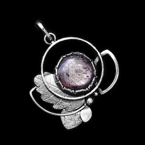 Peut inclure: Pendentif en argent avec une grosse pierre ronde irisée au centre. Le pendentif présente un design stylisé avec des éléments de feuilles et de cercles. La pierre est un mélange de violet, de gris et d'argent. Le pendentif est suspendu à une boucle argentée.