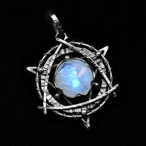 May include: A silver pendant featuring a large, round moonstone with an iridescent glow. The stone is set within a complex geometric design of silver wire and metal shapes, including triangles and circles. The pendant hangs from a silver bail against a black backdrop.