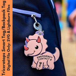 May include: A pink triceratops smart tag with a silver clasp attached to a navy blue backpack. The tag has a friendly face and the words "EMERGENCY CONTACT #" and "nfc" printed on it. The text "Triceratops Smart Tag/Backpack Tag" is on the left.