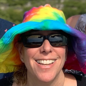 May include: A woman wearing a rainbow tie-dye sun hat with black sunglasses. The hat has a wide brim and is made of a soft, comfortable fabric.
