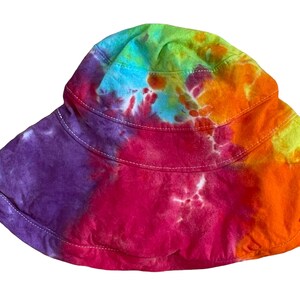 May include: A colourful tie-dye bucket hat with a wide brim. The hat is made of a soft, comfortable fabric and features a variety of colours, including purple, pink, orange, yellow, and green.