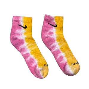 May include: A pair of pink and yellow tie-dye ankle socks with a black Nike swoosh logo on each sock. The socks have a white background with pink and yellow tie-dye patterns.