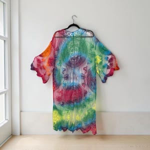 May include: A colorful, flowing kimono-style robe with a vibrant tie-dye pattern. The robe features a spectrum of colors, including red, orange, yellow, green, and blue, with intricate circular designs. The sleeves have a scalloped edge, and the robe hangs on a black hanger.