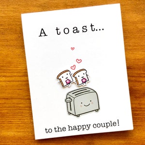 May include: A white card with the text "A toast..." and a cartoon illustration of a toaster with two slices of bread popping up. The bread slices have faces and are holding pink flowers. The card also has the text "to the happy couple!"