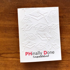 May include: White card with a pattern of graduation caps in raised relief. The text "PHinally Done" is in red and black letters with the word "PH" in red. The text "Congratulations!" is in black letters below.