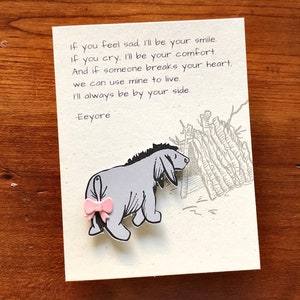May include: A card with a quote from Eeyore, a character from Winnie the Pooh, that reads "If you feel sad, I'll be your smile. If you cry, I'll be your comfort. And if someone breaks your heart, we can use mine to live. I'll always be by your side. -Eeyore". The card features a cartoon illustration of Eeyore with a pink bow.