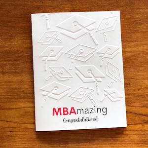 May include: White card with a red and black text that says "MBAmazing Congratulations!" The card is decorated with a pattern of white graduation caps.