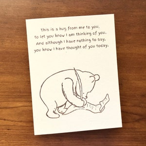 May include: A card with a hand-drawn illustration of a bear writing on a piece of paper. The text on the card reads: "This is a hug from me to you, to let you know I am thinking of you. And although I have nothing to say, you know I have thought of you today."