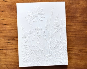 May include: A rectangular white greeting card featuring a raised, embossed design of various wildflowers, including daisies, ferns, and tall grasses, creating a textured, monochromatic botanical pattern across the front surface.