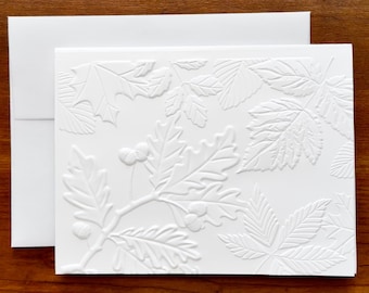 May include: A white rectangular greeting card and matching envelope featuring a raised, embossed design of various leaves and small acorns across the front surface. The texture is monochromatic, highlighting the botanical patterns.