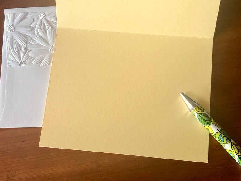 May include: An open, pale yellow greeting card with a patterned pen on the right. A white envelope with embossed daisy-like flowers is on the left. The card and envelope are on a dark wood surface.