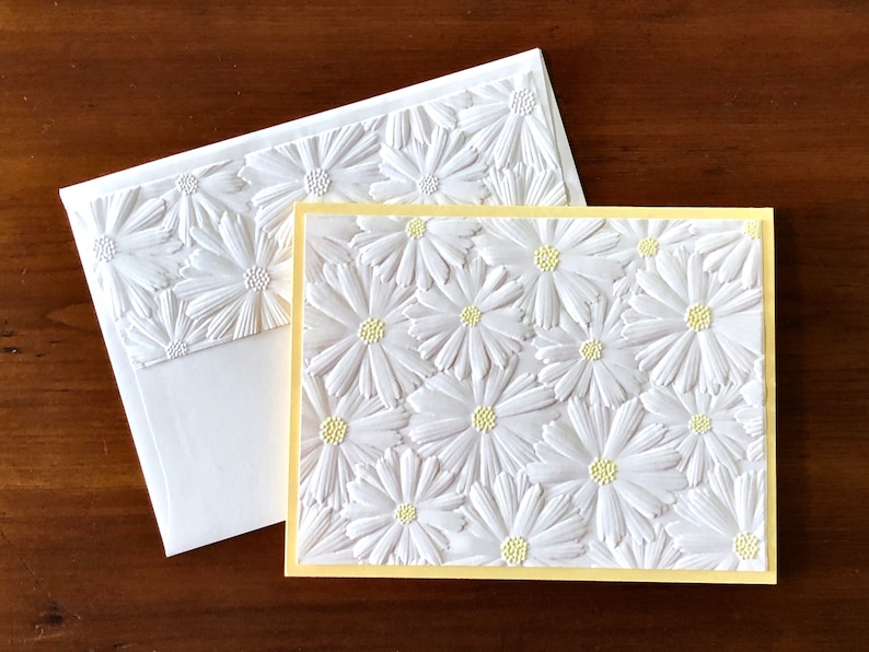 May include: A white greeting card and envelope set with a raised daisy pattern. The card has a yellow border, and the embossed daisies provide a textured surface. The envelope also features the daisy design.