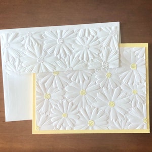 May include: A white embossed daisy stationery set. The set includes a card with a yellow border, an envelope, and a smaller card, all featuring a raised daisy pattern. The daisies have yellow centres. The stationery is on a brown surface.