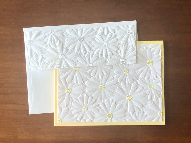 May include: A white embossed daisy stationery set. The set includes a card with a yellow border, an envelope, and a smaller card, all featuring a raised daisy pattern. The daisies have yellow centres. The stationery is on a brown surface.