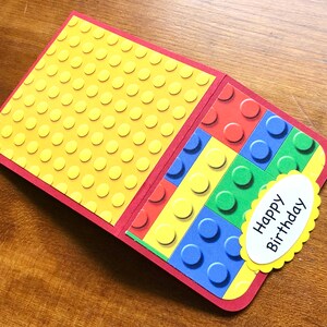 May include: A handmade birthday card with a red border and a yellow interior patterned with raised circles. The right side features a colorful block design, and a yellow scalloped oval with "Happy Birthday" is attached.
