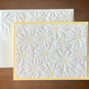 May include: A white embossed greeting card and envelope set with a daisy pattern. The card has a pale yellow border and the daisies have yellow centres. The envelope also has a daisy design.