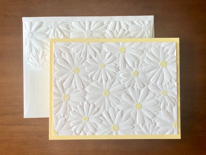 May include: A white embossed greeting card and envelope set with a daisy pattern. The card has a pale yellow border and the daisies have yellow centres. The envelope also has a daisy design.
