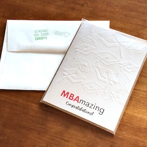 May include: A white greeting card with embossed graduation caps and the words "MBAmazing Congratulations!" in red and black. An accompanying white envelope has the text "Sending you good caroma" and a green heart.