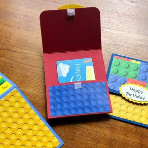 May include: A red gift card holder with a blue textured pocket and a gift card inside. Two additional cards are visible, one with a yellow and blue design and the other with a green and blue design. The words "Happy Birthday" are on a yellow oval.