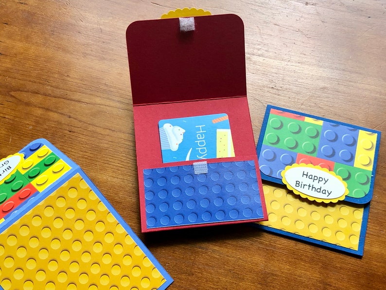 May include: A red gift card holder with a blue textured pocket and a gift card inside. Two additional cards are visible, one with a yellow and blue design and the other with a green and blue design. The words "Happy Birthday" are on a yellow oval.