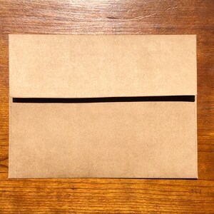 May include: A closed, rectangular kraft paper envelope with a black horizontal band across the middle. The envelope is lying on a wooden surface with a visible wood grain.