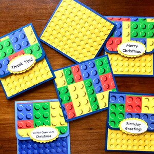 May include: A collection of colorful greeting cards with a Lego brick design. The cards feature blue, green, red, and yellow blocks, with messages like "Thank You!" and "Merry Christmas" on them. The cards are on a wooden surface.