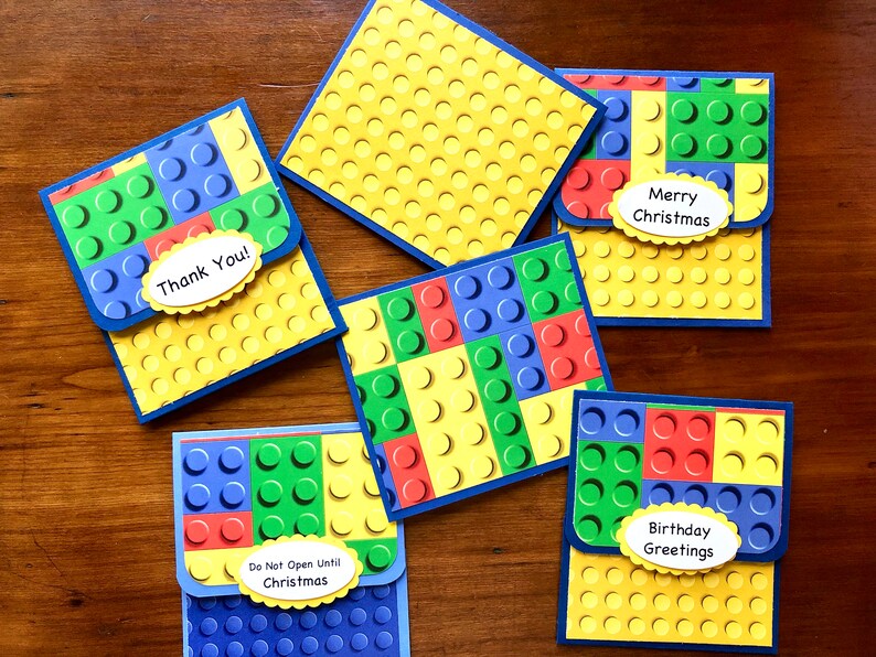 May include: A collection of colorful greeting cards with a Lego brick design. The cards feature blue, green, red, and yellow blocks, with messages like "Thank You!" and "Merry Christmas" on them. The cards are on a wooden surface.
