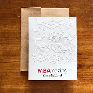May include: White graduation card with embossed mortarboards and the text "MBAmazing Congratulations!" in red and black. The card is inside a brown envelope, set against a wooden background. A perfect gift for a graduate.