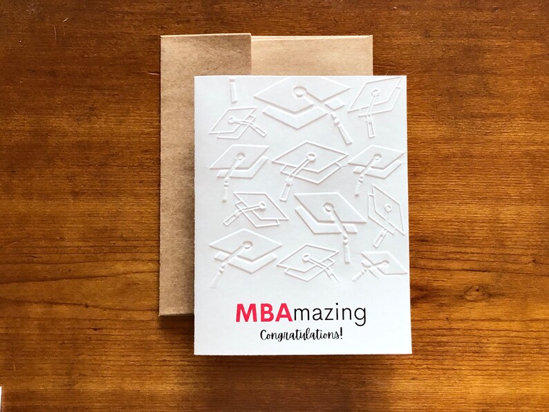 May include: White graduation card with embossed mortarboards and the text "MBAmazing Congratulations!" in red and black. The card is inside a brown envelope, set against a wooden background. A perfect gift for a graduate.