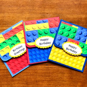 May include: Three handmade birthday cards with a colorful Lego brick design. Each card features a different color scheme: red, blue, and yellow. The cards have the text "Birthday Greetings" and "Happy Birthday" on them.