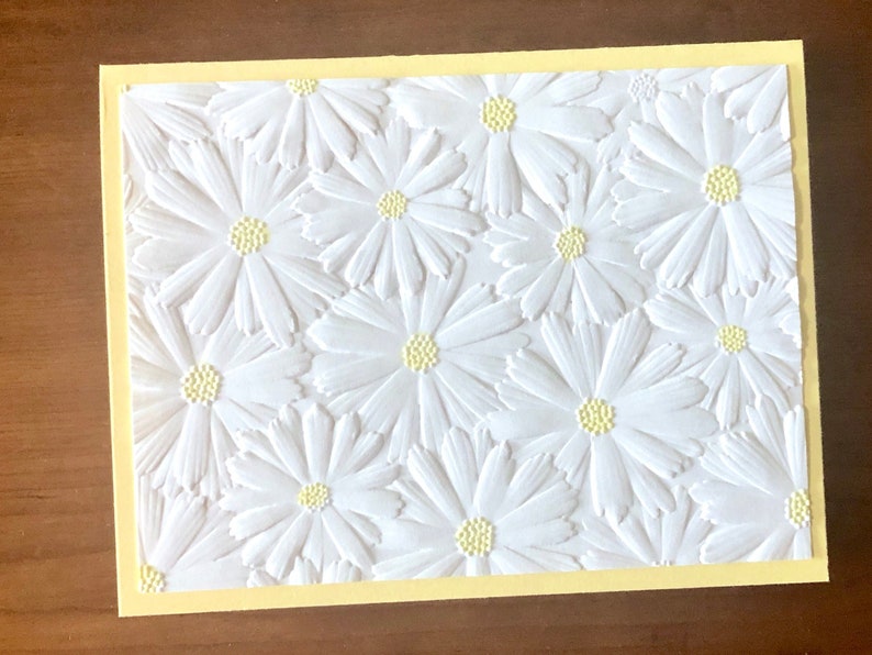 May include: A white card featuring a raised daisy design. The daisies have white petals and yellow centres. The card is set within a yellow border, creating a bright and cheerful design. Ideal for stationery and crafts.