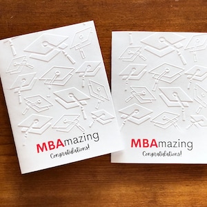 May include: Two white graduation cards with embossed mortarboard designs. The cards have the text "MBAmazing Congratulations!" in red and black. The cards are on a wooden surface.