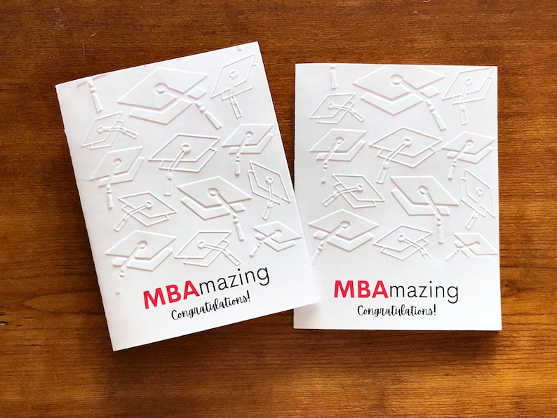 May include: Two white graduation cards with embossed mortarboard designs. The cards have the text "MBAmazing Congratulations!" in red and black. The cards are on a wooden surface.