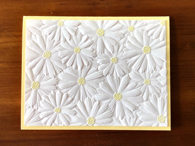 May include: A handmade greeting card featuring a textured white daisy pattern on a white background. The daisies have yellow centres and are set against a pale yellow border. The card is a floral design.