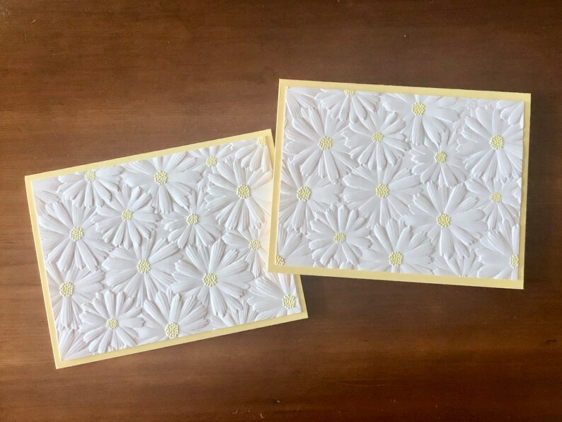 May include: Two rectangular greeting cards featuring a white embossed daisy design on a pale yellow background. The daisy petals are raised, creating a textured effect. The cards are set against a dark wood surface.