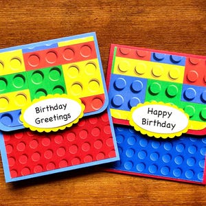 May include: Two birthday cards with a Lego theme. The cards are made of red, blue, yellow, and green paper. The cards have a Lego brick pattern on the front and a white oval with the text 'Birthday Greetings' and 'Happy Birthday' on the front.