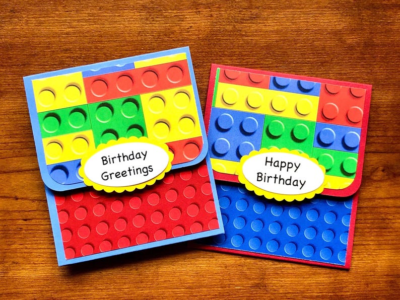 May include: Two birthday cards with a Lego theme. The cards are made of red, blue, yellow, and green paper. The cards have a Lego brick pattern on the front and a white oval with the text 'Birthday Greetings' and 'Happy Birthday' on the front.