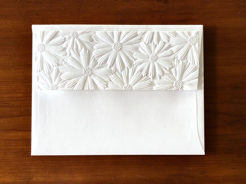 May include: A white envelope with a textured floral design. The top flap features a repeating pattern of embossed daisies. The envelope is made of thick, high-quality paper, suitable for invitations or special notes.
