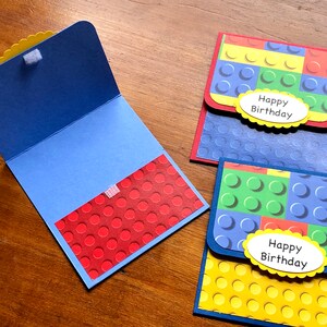 May include: Handmade birthday cards with a colorful Lego brick design. The cards feature blue, red, green, and yellow blocks, with a white oval that says "Happy Birthday". One card is open, revealing a red interior with a dot pattern.