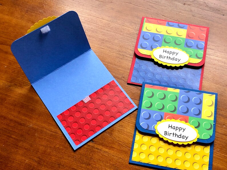 May include: Handmade birthday cards with a colorful Lego brick design. The cards feature blue, red, green, and yellow blocks, with a white oval that says "Happy Birthday". One card is open, revealing a red interior with a dot pattern.