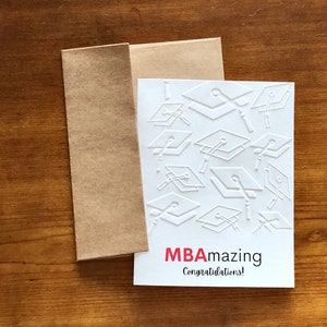 May include: A white MBA graduation card with embossed mortarboards and the words "MBAmazing Congratulations!" in red and black. The card is paired with a brown envelope, set against a wooden background.