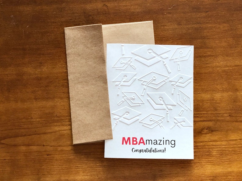 May include: A white MBA graduation card with embossed mortarboards and the words "MBAmazing Congratulations!" in red and black. The card is paired with a brown envelope, set against a wooden background.
