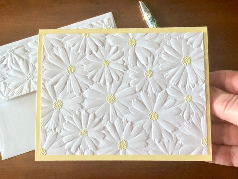 May include: A handmade greeting card with a yellow border and a white embossed daisy pattern. An envelope with a matching daisy design is visible. A pen with a floral design is in the background.