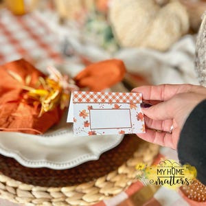 May include: A fall-themed place card with a gingham top border and a white center for writing. The card is decorated with autumn leaves in shades of orange and red. The card is held over a white plate and a woven placemat.
