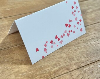 Puede incluir: Tarjeta de lugar blanca con un diseño de corazones rosas. Los corazones están dispersos por la tarjeta, con una mayor concentración en el lado derecho.