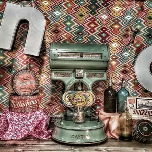 May include: A vintage green scale with a Dayton logo, surrounded by various items including candy, old-fashioned bottles, and a box of Snickers candy bars. The scale is on a wooden surface with a patterned rug in the background.