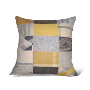 May include: A square cushion with a patchwork design featuring various shades of grey, yellow, and brown. The cushion is made of a soft, textured fabric.