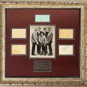 May include: A framed display featuring a black and white photograph of The Rat Pack from the 1960 film Ocean's Eleven, surrounded by five individual autographs on cream paper and a central plaque with the text The Rat Pack, Frank Sinatra, Dean Martin, Sammy Davis Jr., Peter Lawford, Joey Bishop, in Warner Brothers Ocean's Eleven 1960, all set within a decorative gold frame.